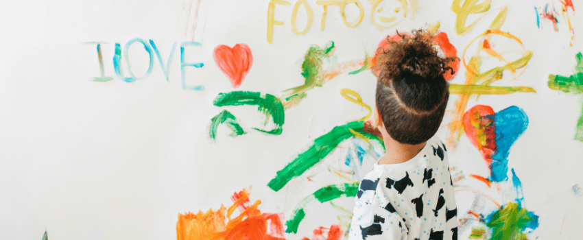 A young child paints words on a wall with supplies their teacher bought using discounts.